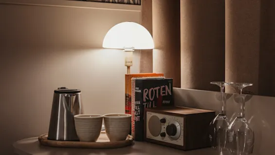 Ett varmt upplyst nattduksbord med en vattenkokare, koppar, böcker inklusive ROTEN TILL ROMAN, en radio och vinglas.