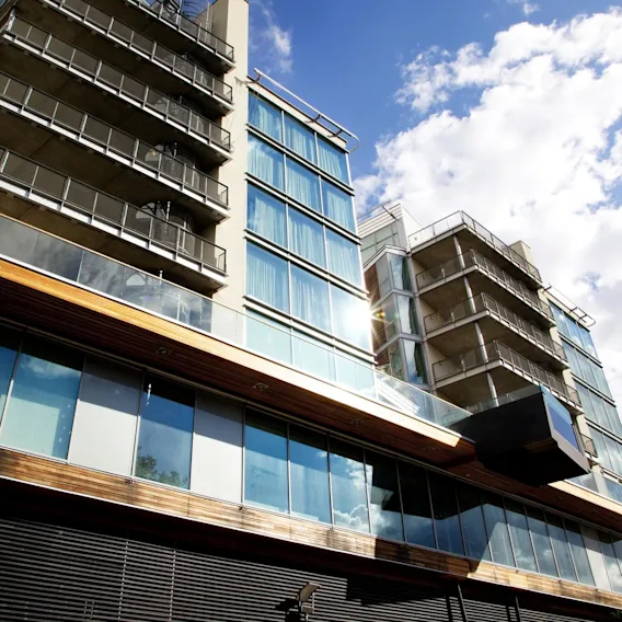A modern glass and steel building rises with multiple balconies, set against a partly cloudy blue sky. The facade reflects sunlight, emphasizing its contemporary architectural style.