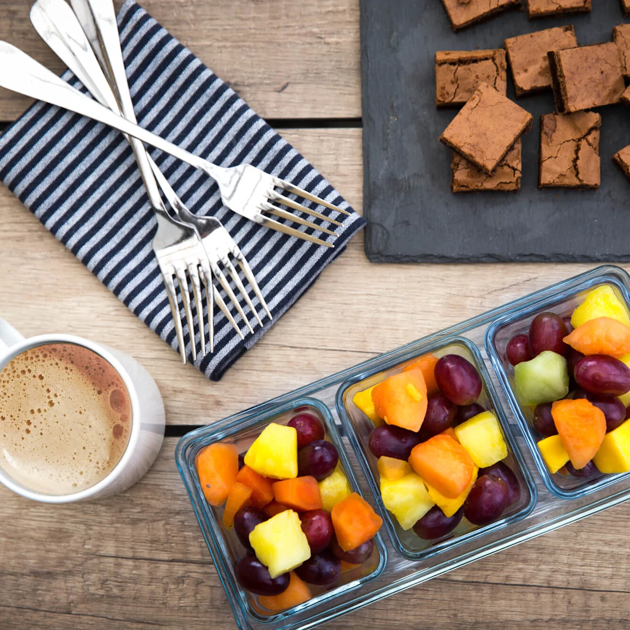 Coffee cup beside glass containers of fruit salad and brownies on wooden table.