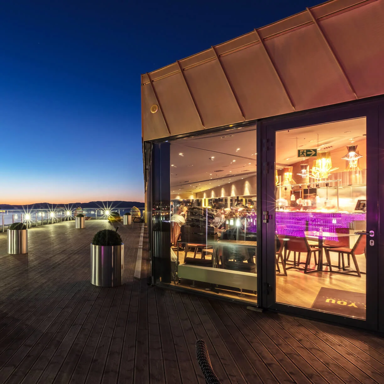 Rooftop terrace with metallic planters holding shrubs, overlooking water and mountains at dusk; adjacent to a glass-front restaurant illuminated with warm lights and a purple bar counter.
