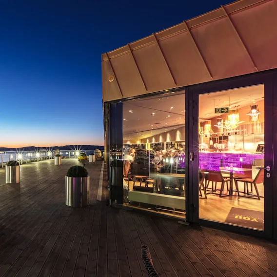 Rooftop terrace med metallplantere med busker, med utsikt over vann og fjell i skumringen; ved siden av en glassfrontet restaurant opplyst med varme lys og en lilla bardisk.
