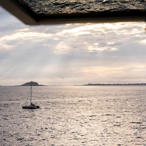 En seilbåt driver på et glitrende hav under en skyete himmel, synlig gjennom et vindu. En fjern øy ligger i horisonten.