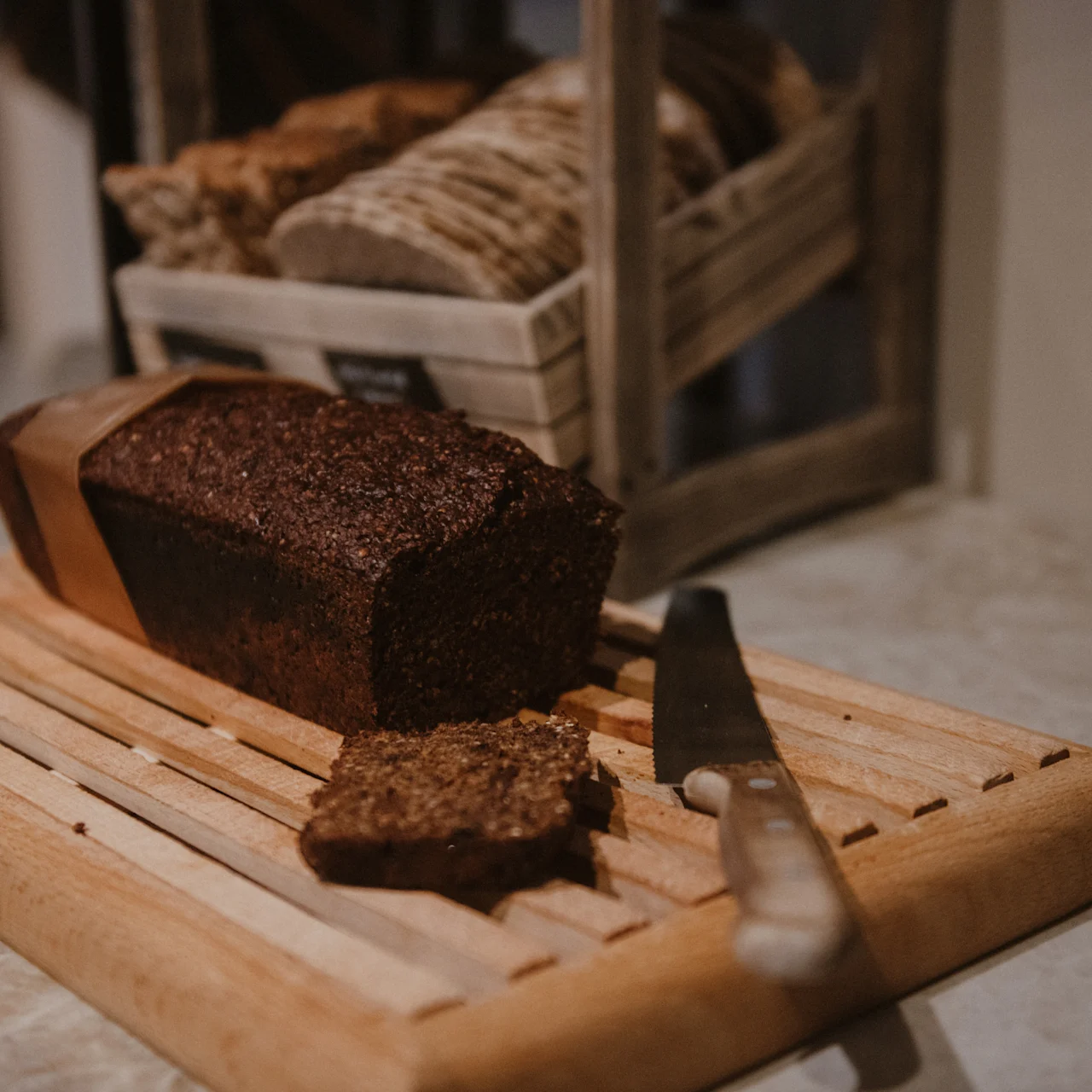 A loaf of dark bread sits sliced on a wooden cutting board, accompanied by a knife. In the background, a basket contains additional loaves.