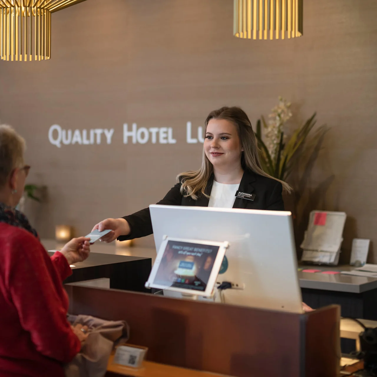 Receptionist ger ett kort till en gäst vid hotellets incheckningsdisk. Elegant belysning ovan, blommor i närheten. Skylten lyder "QUALITY HOTEL LLE.” Receptionen känns modern och välkomnande.