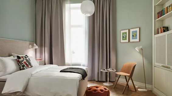 En seng med hvitt sengetøy står mot en mintgrønn vegg, omgitt av tremøbler, en liten oransje puff, to innrammede kunstverk og myk belysning fra et vindu.