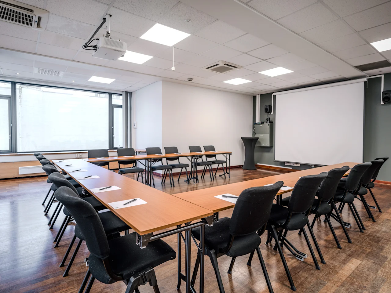 Moderni kokoushuone U-muotoisella pöytäjärjestelyllä, projektorilla ja näytöllä, valmiina esitykseen tai kokoukseen.