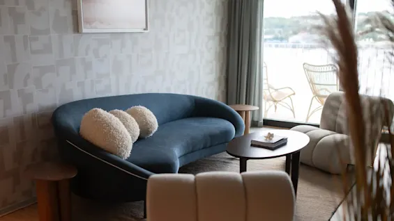 A blue sofa with three fluffy pillows sits in a modern living room. Nearby, a coffee table holds a book. Large windows reveal a balcony with wicker chairs.
