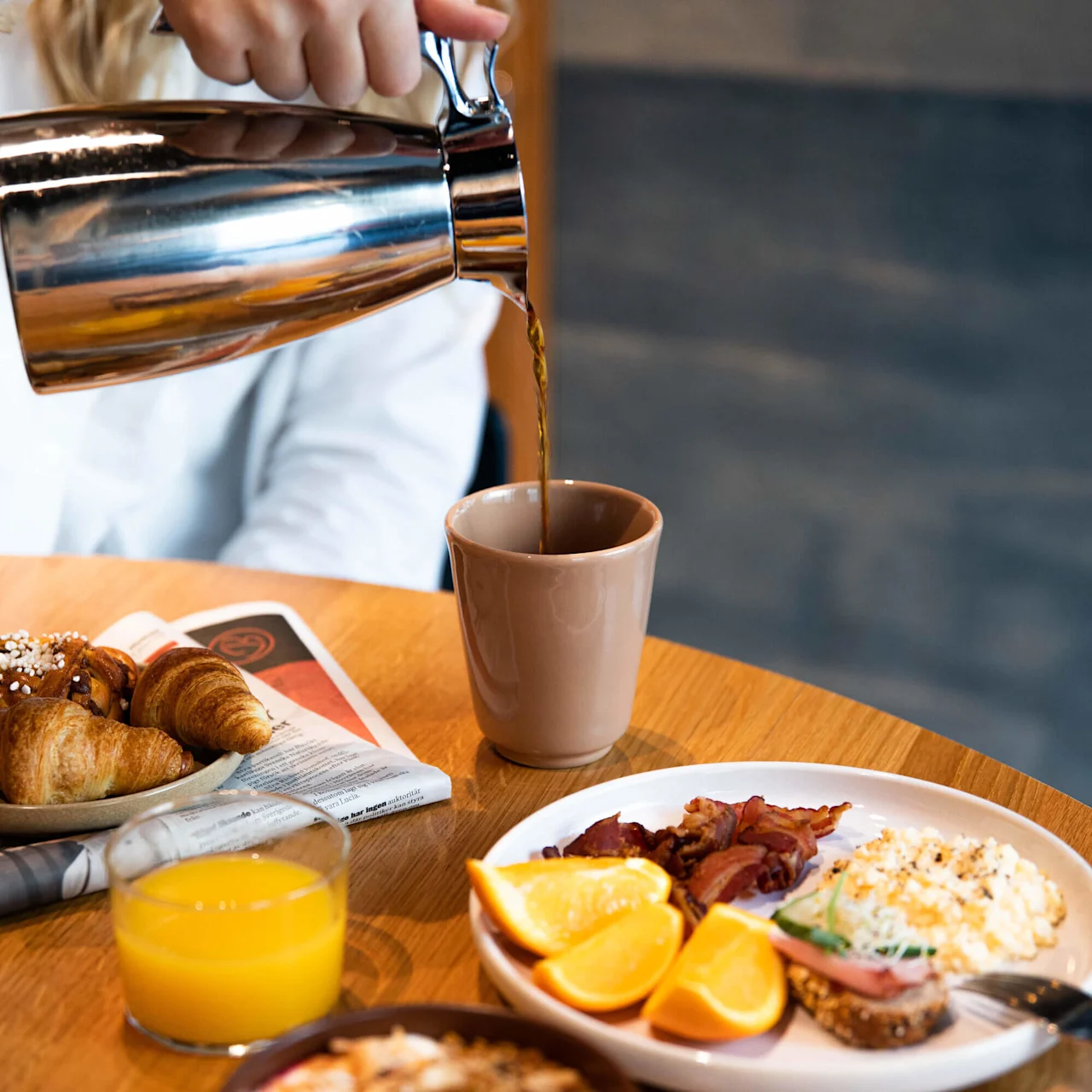 A hand pours coffee from a silver pot into a mug on a table. Nearby, a plate with croissants, a newspaper, orange juice, and breakfast items suggests a morning setting.