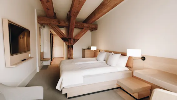 Ett ljust och modernt hotellrum med en stor säng, synliga takbjälkar i trä och minimalistisk ljus inredning.