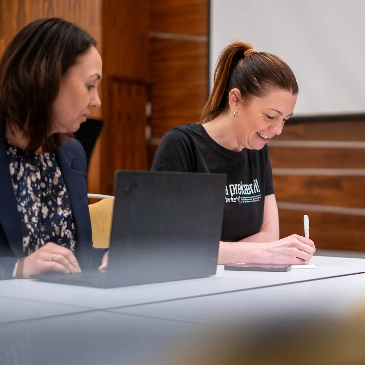 A woman is writing with a pen in a meeting room, smiling, surrounded by others taking notes. Text on her shirt partially reads: "a prekaer". 