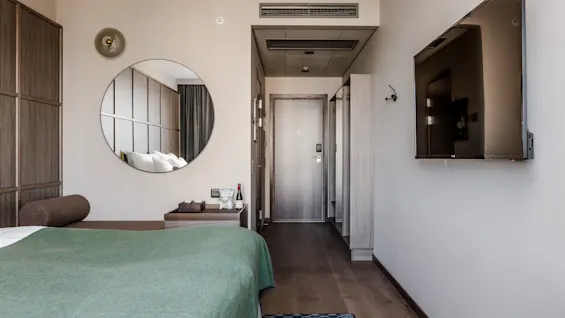 En seng med en brun bolster og grøn teppe står i et moderne hotellrom. Trepanel, en veggmontert TV og et stort rundt speil skaper en minimalistisk atmosfære.