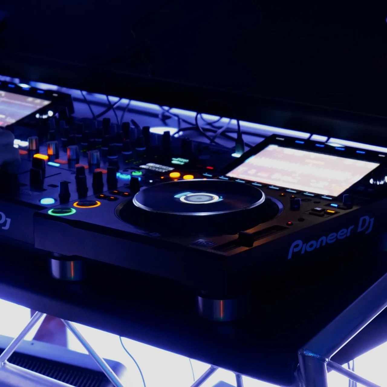 DJ-udstyr er i brug med farverige knapper og skærme oplyst i et mørkt miljø. Enheden er mærket "Pioneer DJ".