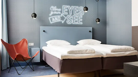 Et moderne soverom med to enkeltsenger under hengende lamper, en skinnstol og en vegg med teksten "Close your EYES AND SEE." Rommet har et minimalistisk design.