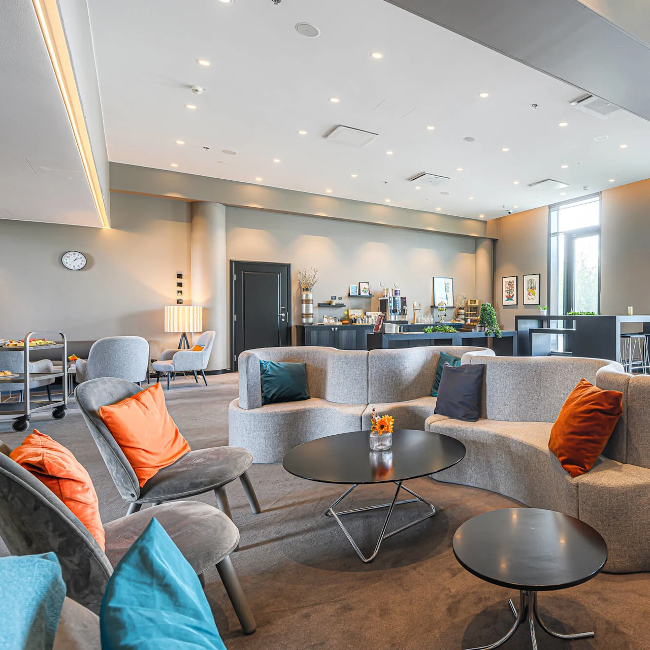 A modern lounge features curved gray sofas with colorful cushions, low black tables, and a windowed wall providing natural light. A clock and refreshments are visible on the counter.