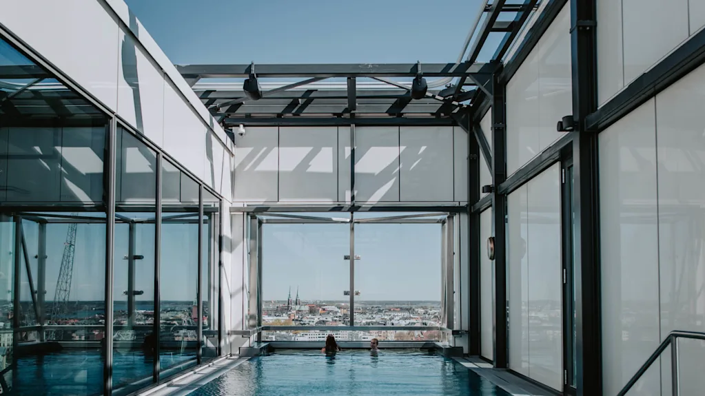 Två personer i en inomhuspool slappnar av, omgivna av glasväggar och metallbalkar, med en stadsbild synlig i bakgrunden. En "1.2 m" djupskylt finns vid poolkanten.