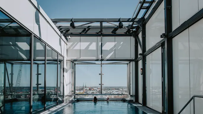 To personer i et innendørs basseng slapper av, omgitt av glassvegger og metallbjelker, med en byutsikt i bakgrunnen. Et "1.2 m" dybdeskilt er på bassengkanten.