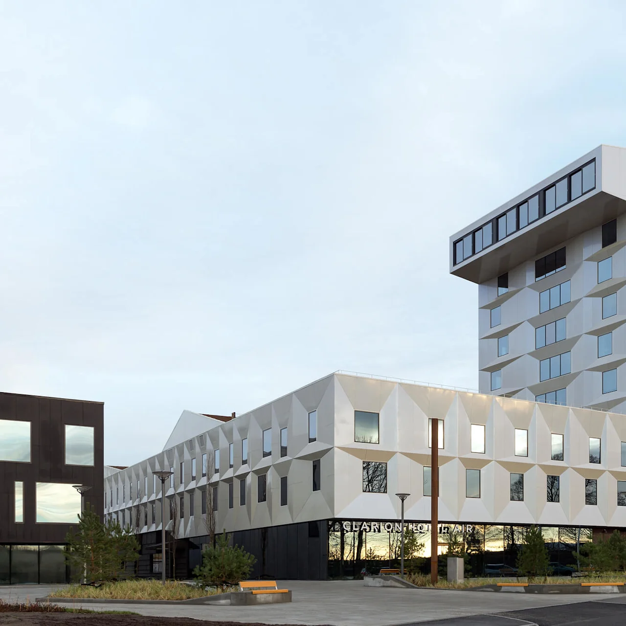 En moderne, geometrisk designet bygning med vinklede vinduer og hvit fasade står høyt. Nedenfor står teksten "CLARION HOTEL AIR." I nærheten flyr et fly i en delvis skyet himmel.