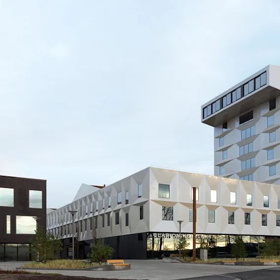 En moderne, geometrisk designet bygning med vinklede vinduer og hvit fasade står høyt. Nedenfor står teksten "CLARION HOTEL AIR." I nærheten flyr et fly i en delvis skyet himmel.