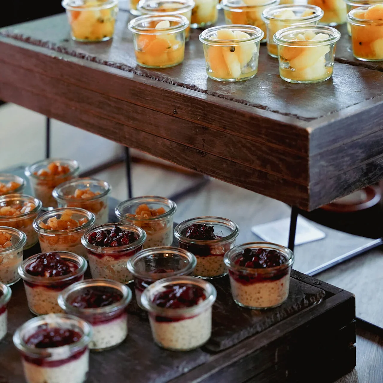 Flere små dessertkopper på en trappeformet træholder, der viser forskellige toppings, ved en cateringbegivenhed.