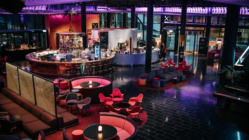 A circular bar with stools accommodates patrons in a modern lounge with colorful chairs and sofas. Soft purple and blue lighting creates a vibrant, inviting atmosphere.