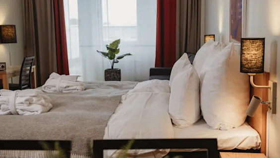 A neatly made bed with fluffy pillows is situated in a cozy hotel room. Soft lighting and curtains frame a window, while a potted plant adds greenery on a table.