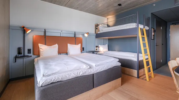 Et hotellrom har en dobbeltseng og en køyeseng med gul stige, komplementert av moderne belysning og tregulv i et minimalistisk design.