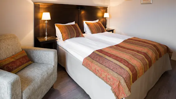 En snyggt bäddad säng med mönstrad överkast och kuddar, ackompanjerad av en stol, i ett varmt belyst hotellrum med konstverk.