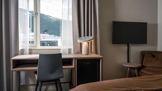A desk with a chair and lamp sits by a window showing a mountain view. Nearby, a TV is mounted on the wall. The room features curtains and a soft bedspread.