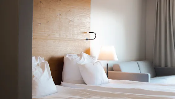 Et hyggeligt hotelværelse med en redt seng, bløde hvide puder, en varm lampe og en komfortabel sofa i baggrunden.