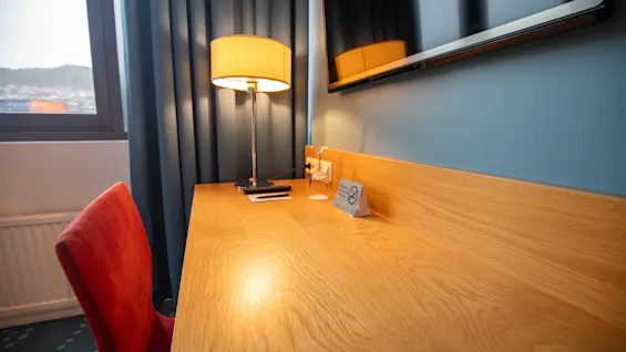 Et skrivebord i tre har en lampe, blokk og et skilt med "No Smoking". En rød stol står i nærheten, under en veggmontert TV, ved siden av et gardinbelagt vindu.