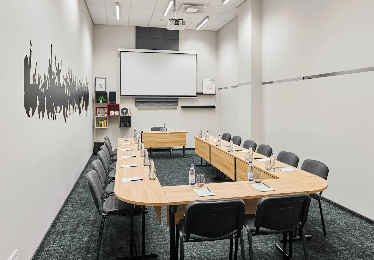 Conference table arranged in a U-shape with water bottles and notepads, surrounded by gray chairs. A projector screen is on the wall, with shelves and wall art nearby.
