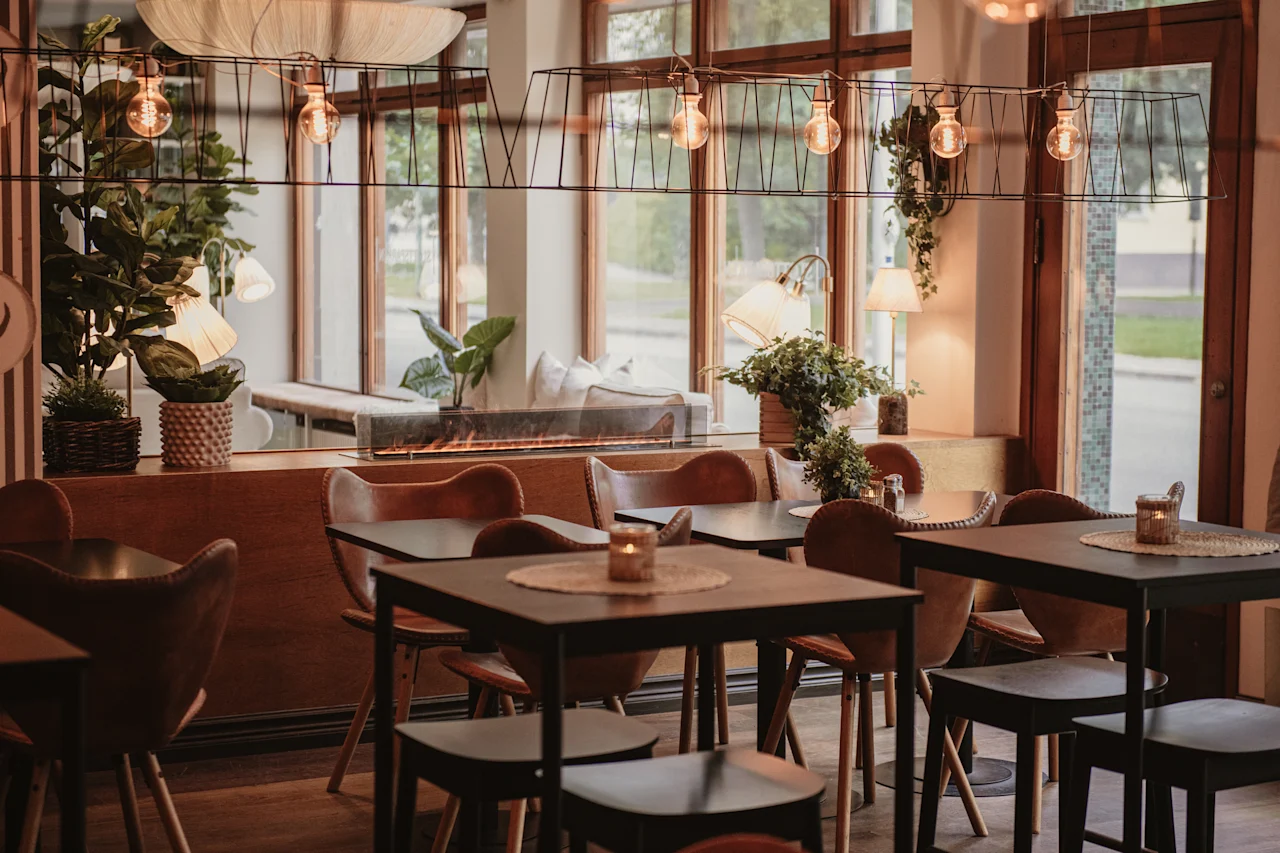 A warm and inviting restaurant interior with tables, chairs, plants, and soft lighting, creating a cozy atmosphere.