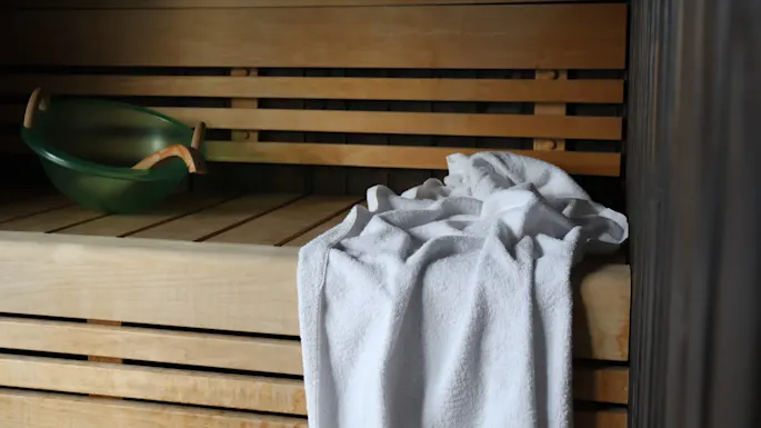 En grønn badstuebøtte med et trehåndtak ved siden av et krøllete hvitt håndkle på trebadstuebenker.