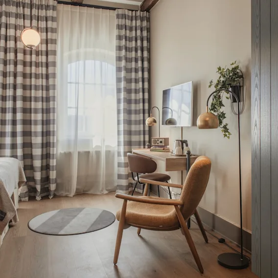 Viihtyisä hotellihuone, jossa sänky, työpöytäalue ja mukava nojatuoli, ruudulliset verhot ja lämmin valaistus.