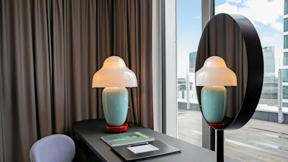 A lamp with a mushroom-shaped shade sits on a desk alongside a chair, papers, and a pen. Its reflection appears in a nearby circular mirror; a window overlooks a cityscape.