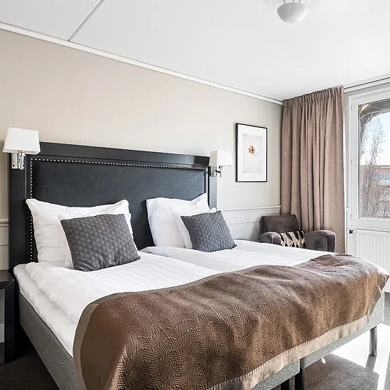 A neatly made bed takes center stage in a well-lit bedroom with windows overlooking buildings.