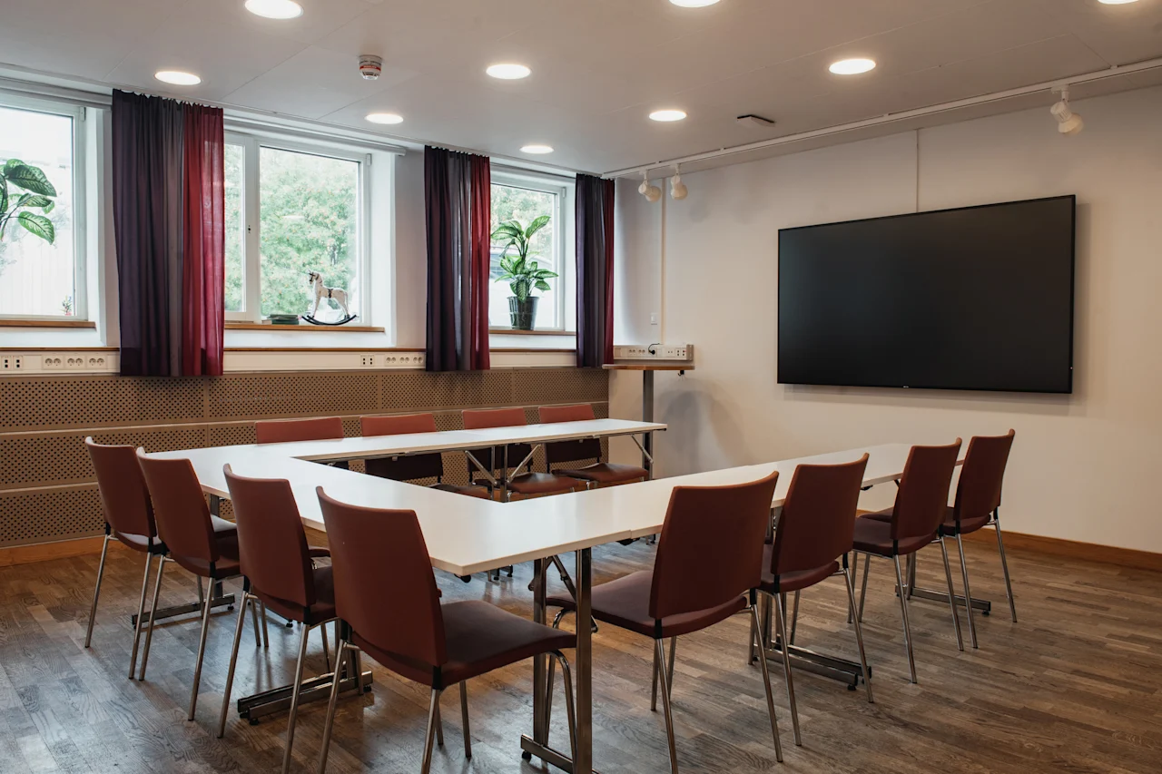 A modern conference room at Home Hotel Uppsala with a U-shaped table, chairs, large windows, and a wall-mounted screen.