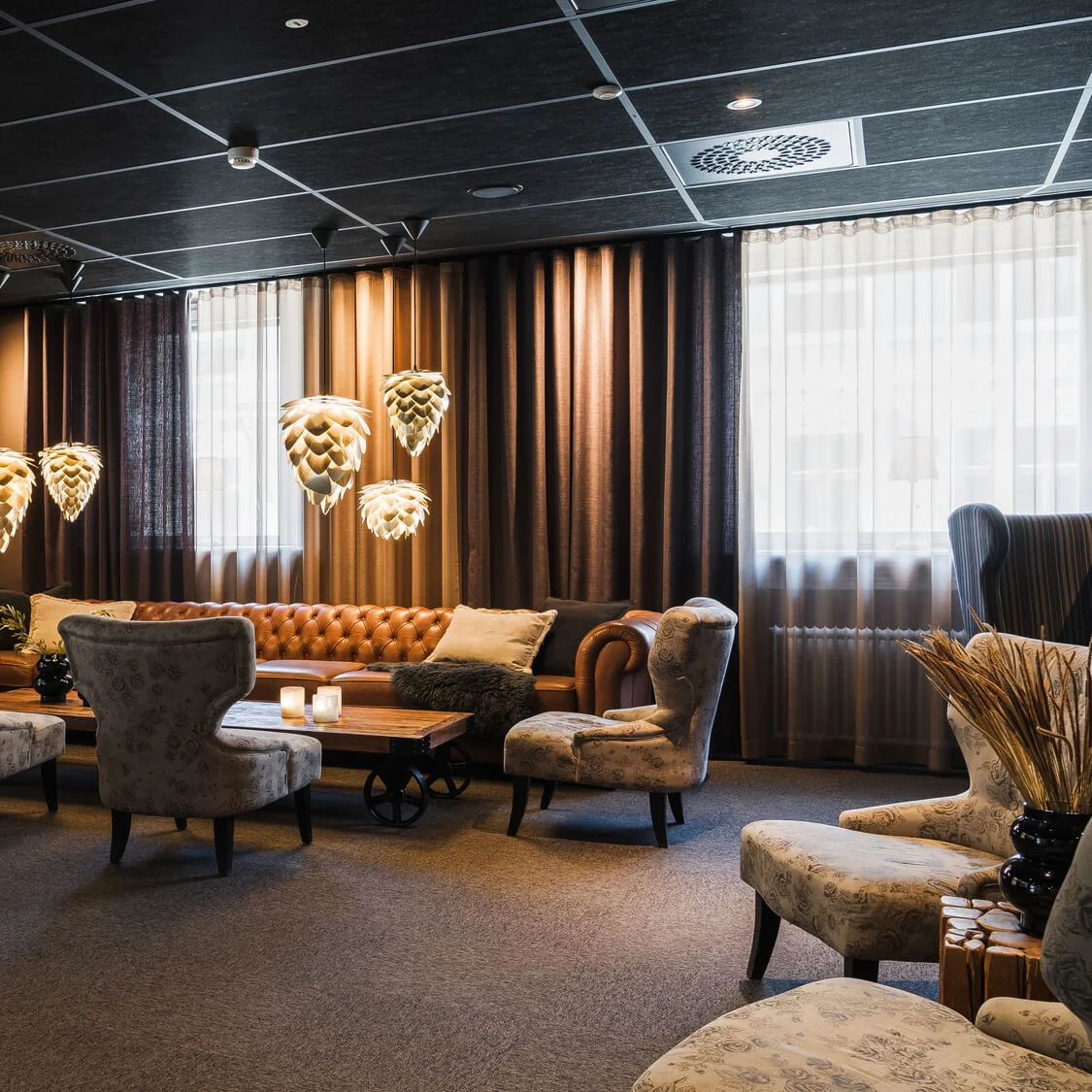 En mysig lounge har en brun lädersoffa belyst av kronärtskocksformade pendellampor. Omgivande fåtöljer och ett soffbord i trä skapar en varm, inbjudande atmosfär med mjuk belysning.