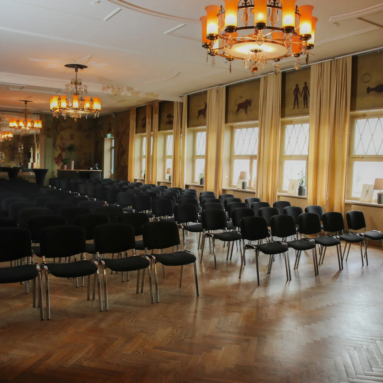 Rows of black chairs are arranged in a large, elegant room with decorative chandeliers and tall windows covered by curtains, creating an atmosphere suitable for a conference or event.