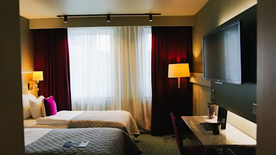 En hotellrum med två sängar med quiltade överkast, ett skrivbord med stol, en väggmonterad TV och ett stort fönster med vinröda gardiner. Belysningen är varm och inbjudande.