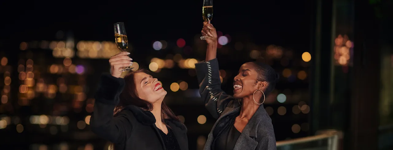 Two people hold champagne glasses aloft in celebration on a balcony, surrounded by city lights at night.