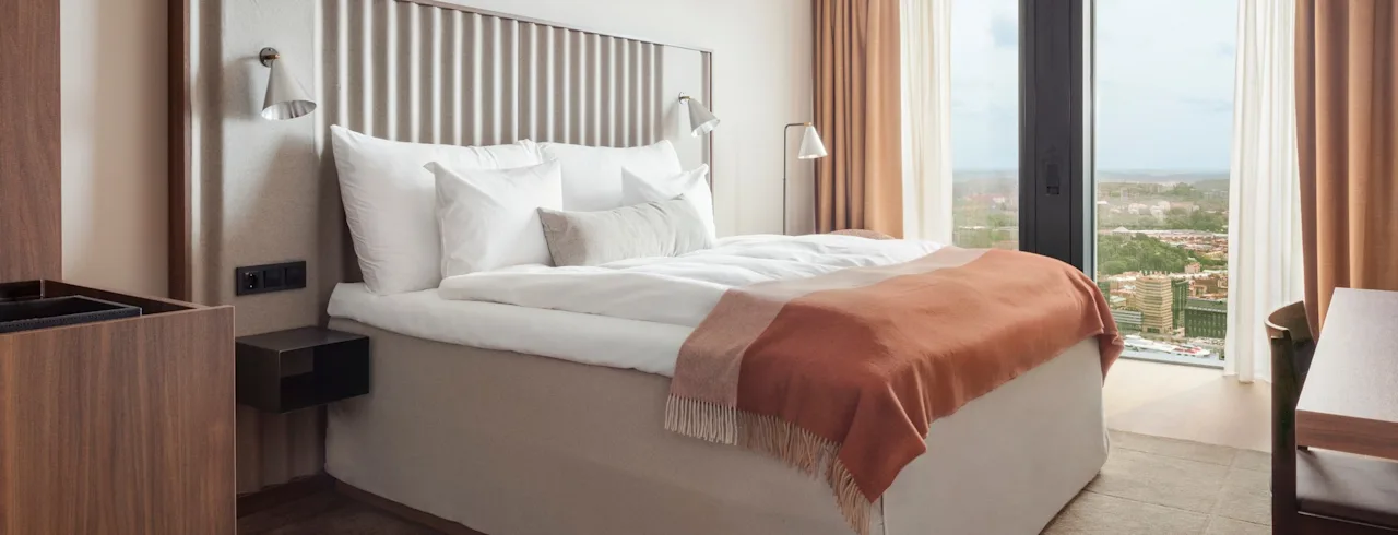 En pænt redt seng med hvidt sengetøj og et orange tæppe i et moderne hotelværelse. Store vinduer giver udsigt over landskabet udenfor.