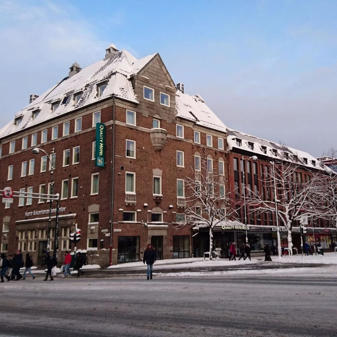Et snedækket murstenshotel står på et travlt gadehjørne, omgivet af fodgængere og snedækkede træer. Skilte lyder "Quality Hotel" og "Kurs & Konferansesenter".