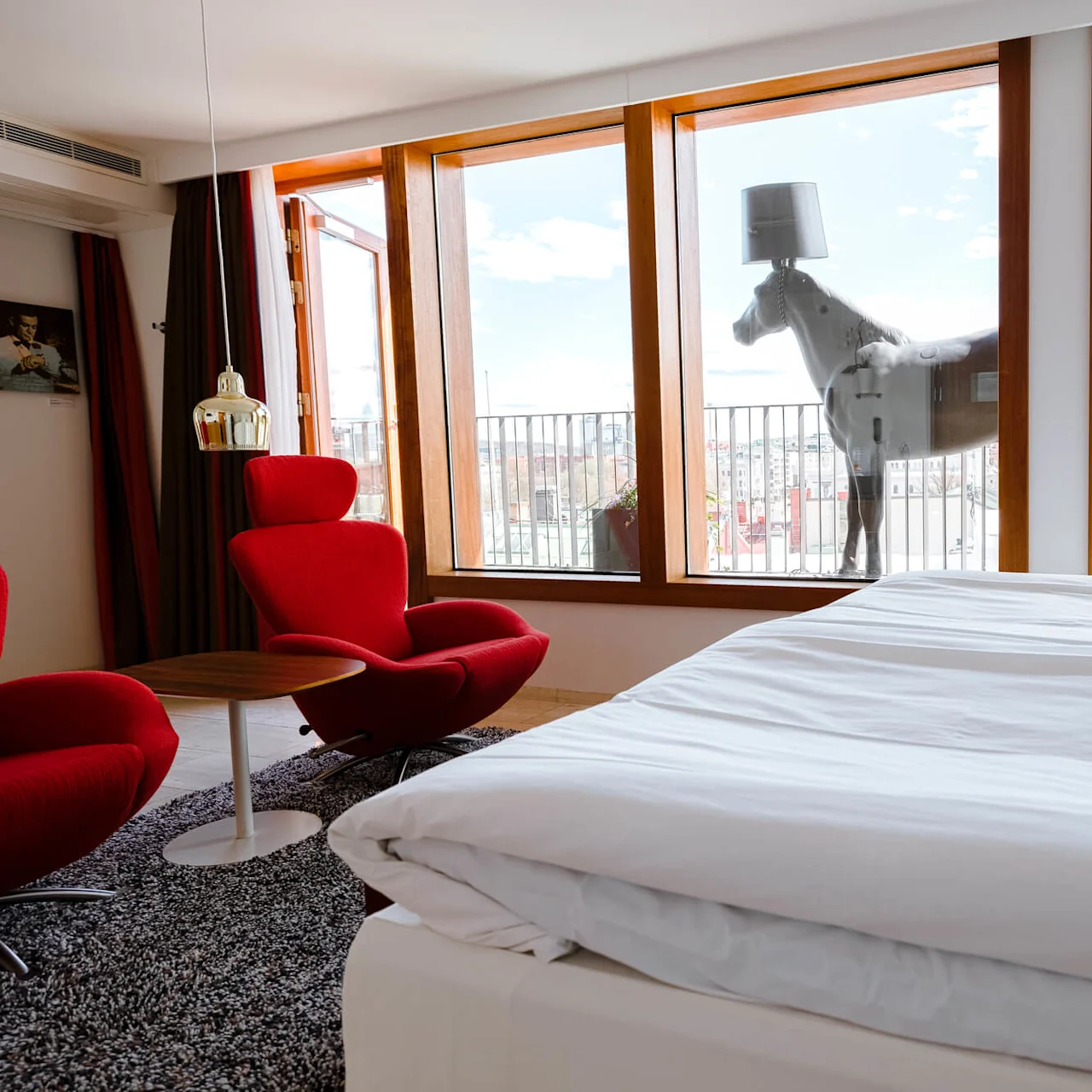 A large bed with white linens is in a modern room. Two red chairs and a small table sit by wide windows overlooking a balcony with a horse statue.
