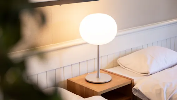 En hvid lampe oplyser et natbord mellem to pænt redte senge i et blødt oplyst rum, med en plante delvist synlig i forgrunden.