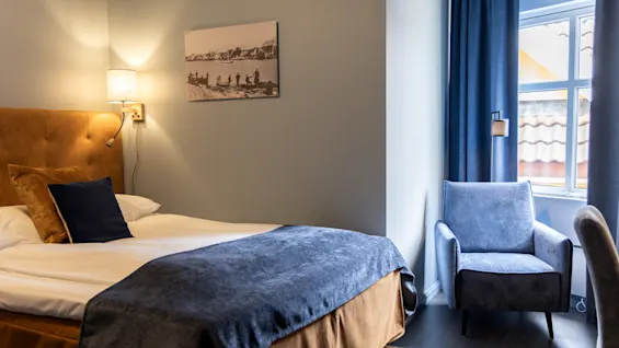 A cozy bedroom features a neatly made bed with blue and brown linens beside a lit wall lamp. A blue armchair sits near a window with closed curtains.