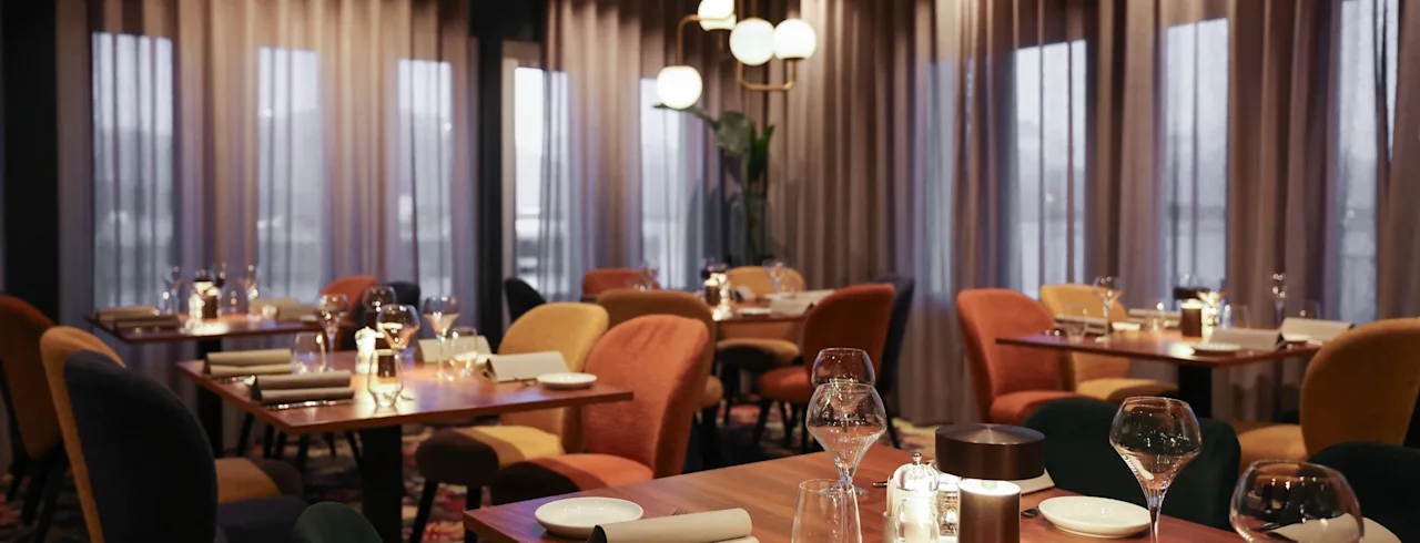 Elegante spisebord er ryddig arrangert, dekket med tallerkener, glass og servietter, under dempet, varm belysning i en moderne restaurant med store vinduer og tynne gardiner.