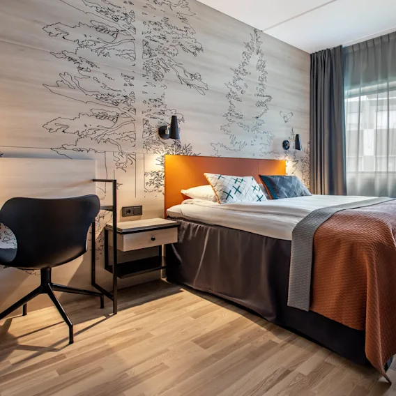 En pent oppredd seng med oransje og grått sengetøy står i et moderne rom med tregulv, kunstneriske veggtegninger og myk belysning fra en stor lampe.
