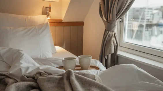 A cozy bedroom with a bed, warm blankets, and two cups on a tray, next to a window with a view.