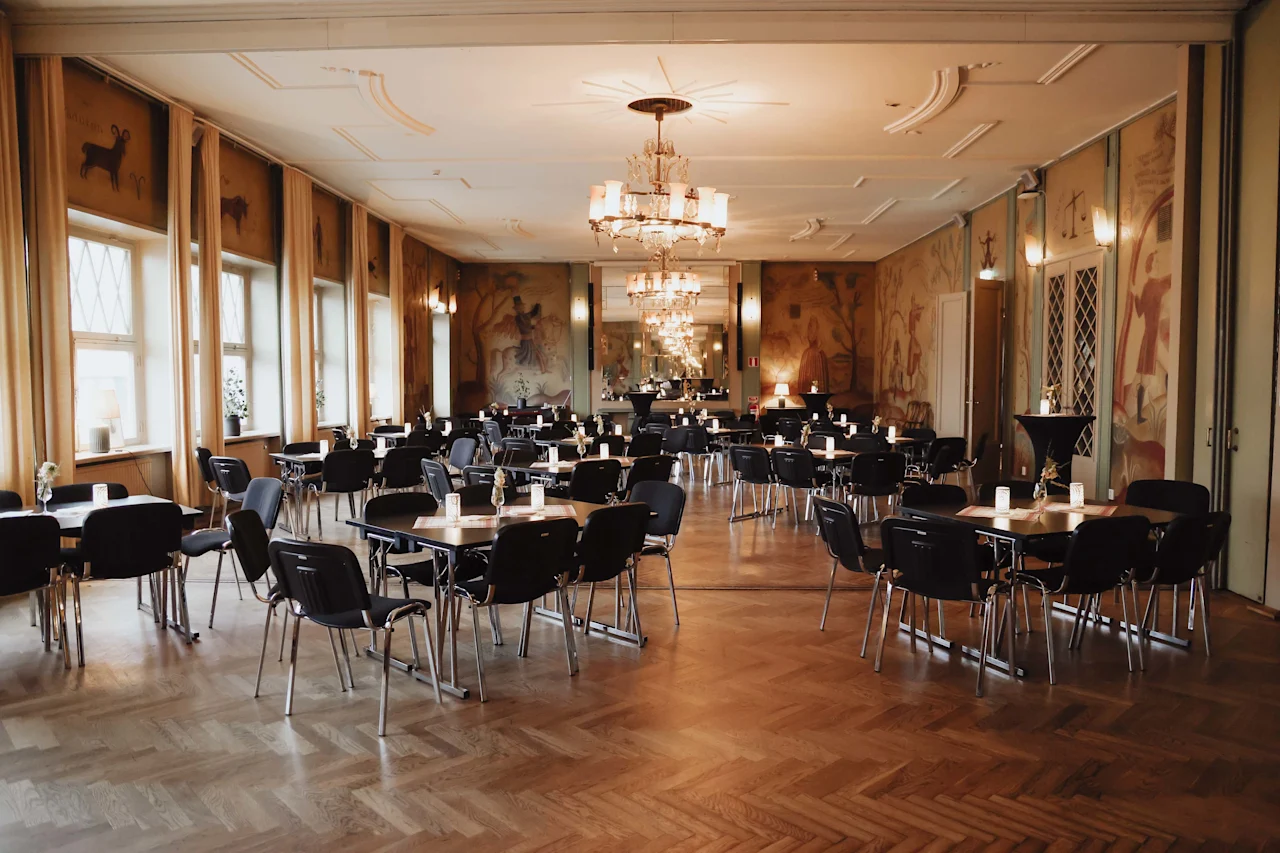Tables and chairs are arranged neatly in an elegant, spacious room with chandeliers and patterned wall art. Soft lighting creates a warm, inviting atmosphere, complemented by large windows.